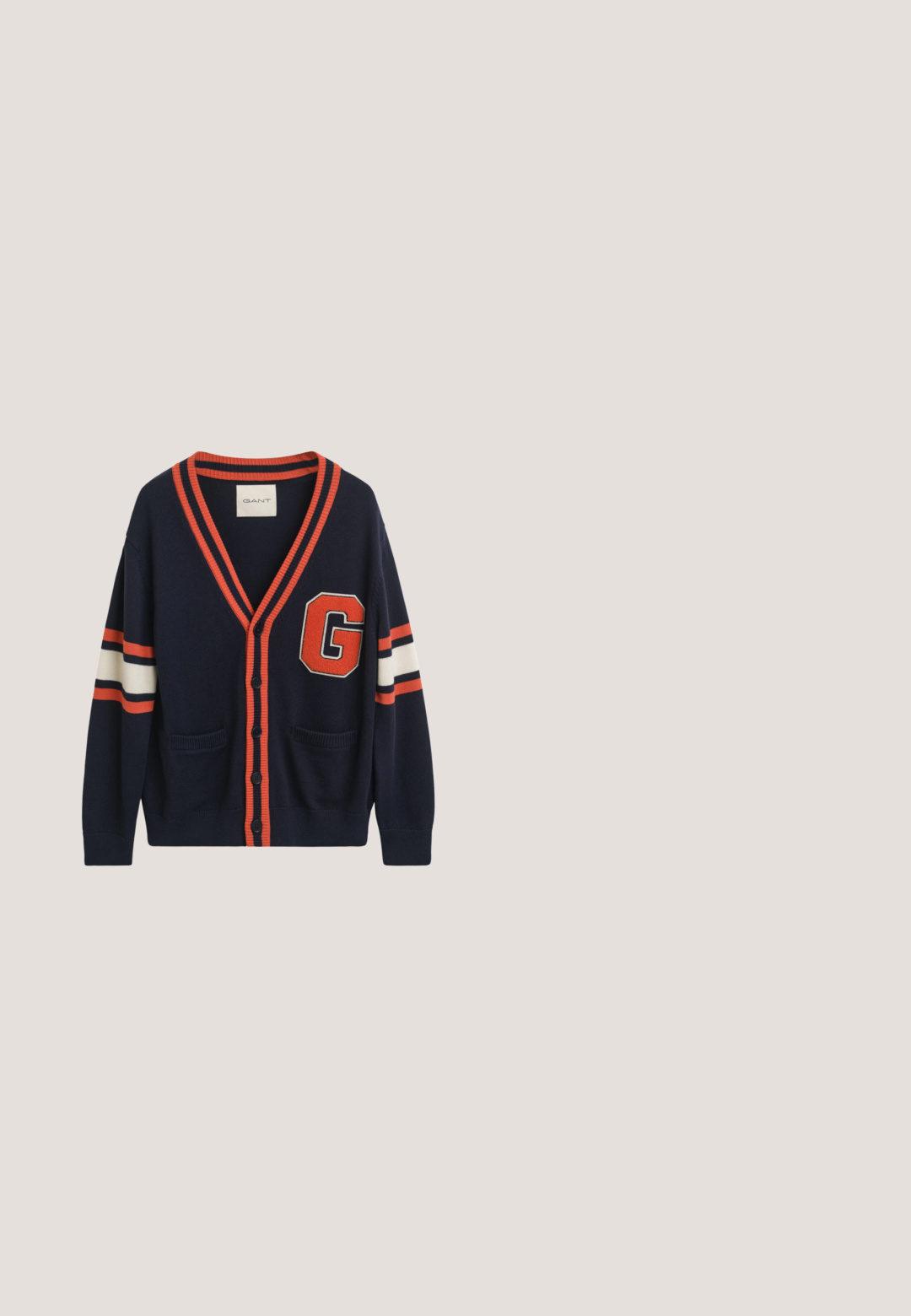 Cárdigan navy hecho de tela de punto, con rayas naranjas y blancas en las mangas, un gran parche con la letra "G" y dos bolsillos delanteros.; Camisa beige de manga larga con botones, cuello y pequeño logo bordado en el pecho izquierdo, mostrada sobre fondo blanco.; Pantalones azul marino de pierna recta con trabillas para cinturón, cierre frontal de botones y bolsillos laterales, mostrados sobre un fondo liso.; Zapato mocasín de ante marrón para hombre flotando sobre una alfombra beige con un sofá beige a juego en el fondo.; Un collar de cadena plateada, un collar de cuentas marrones y negras con cierre de plata, y una pulsera de cuentas marrones y negras de múltiples hilos.; Cinturón beige tejido enrollado con hebilla de metal plateado y ribete de cuero beige sobre un fondo liso y claro.