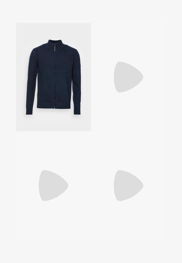 Marineblå zip-up sweater med høj krave, ribbede manchetter og kant, glat strik tekstur og detaljer med sidelapper.; Hvid bomulds T-shirt med korte ærmer, rund halsdesign og en glat tekstur. Etiket synlig ved halsudskæringen. Enkel og klassisk stil.; Mørkeblå denim jeans med lige ben design, lys gylden stikning, forlommer og en enkelt knaplukning i taljen.; Sort sneaker med hvid sål svævende over en beige tekstureret tæppe nær et rundt trætabel med stablede bøger.