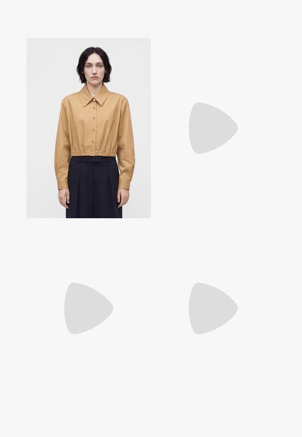 Femme aux cheveux courts et foncés portant une chemise courte boutonnée beige et un pantalon large taille haute foncé, debout devant un fond uni.; Blouse beige sans manches avec encolure drapée et ourlet droit, en tissu lisse, étiquette Tiger of Sweden à l'intérieur du col.; Pantalon beige clair à jambes larges avec taille élastique et cordon, poches latérales et détails subtils de coutures.; Escarpin en daim taupe à talon haut, bout pointu et talon en forme de cône sur fond blanc.