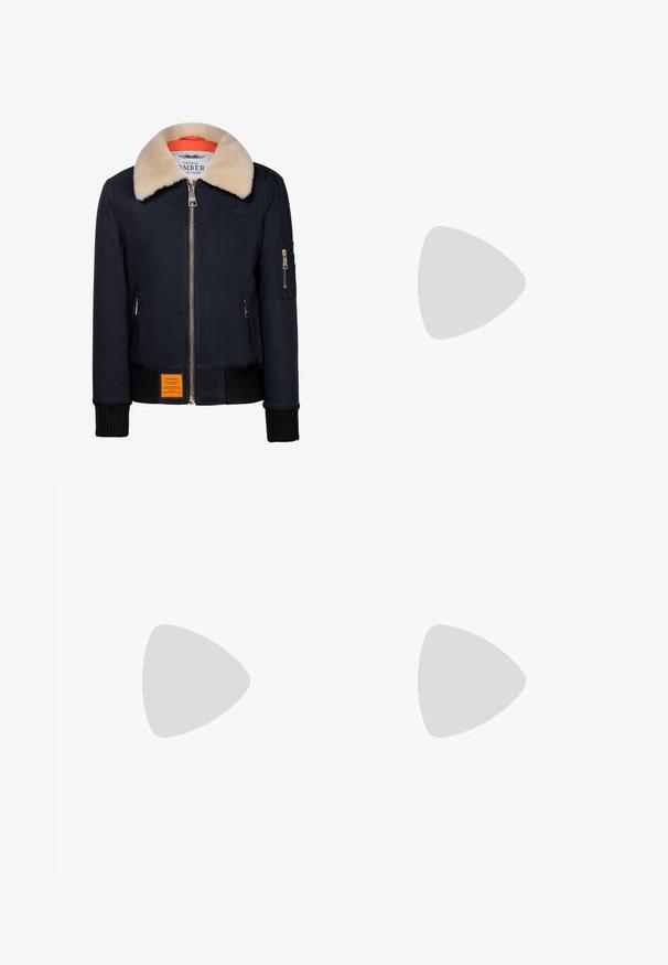 Marine bomberjack met een nepbontkraag, gouden rits, ribgebreide manchetten en een oranje voering. Heeft een oranje label aan de voorkant.; Zwarte geribbelde T-shirt met een hoge hals en korte mouwen. Gladde, aansluitende pasvorm zonder patronen of zichtbare hardwaredetails.; Persoon die zwarte skinny jeans met hoge taille en zwarte puntige pumps draagt, staand tegen een effen witte achtergrond.; Tan suède enkellaarsjes met bruine leren accenten, gouden oogjes en een getextureerde zool. Witte sokken zichtbaar boven de laarsjes.