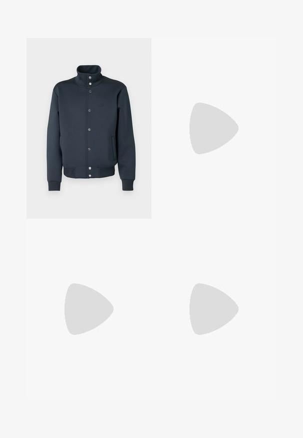 Veste sweat bleu marine avec col montant, fermeture à boutons-pression, poignets en tricot et poches latérales. Présente un logo Lacoste sur la poitrine.; Polo vert à manches courtes avec col, deux boutons et un petit logo crocodile brodé sur la poitrine.; Jogging léger crème avec une taille élastique, des chevilles resserrées, des fentes latérales et un petit logo vert sur la cuisse supérieure.; Baskets noires et beiges avec motifs ondulés, design à lacets et semelle épaisse sur fond clair uni.