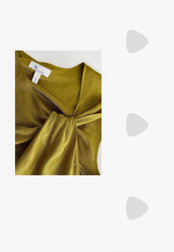 Olivengrøn bluse med en snoet stofdetalje foran, lavet af et let, gennemsigtigt materiale. Har et mærke mærket "N. PREMIUM COLLECTION."; Sorte ruskind højhælede pumps med spids tå, glat tekstur og en slank silhuet. Har en slank stiletto-hæl og åbne sider.; Beige læderhåndtaske med snørebånd og sølvfarvede metaløjer samt en aftagelig sølvkæderem lagt ved siden af.; Guldtonet metalhalskæde med et glat kvindesymbol vedhæng, hvilket har en delikat kæde og en justerbar clasp for længde.