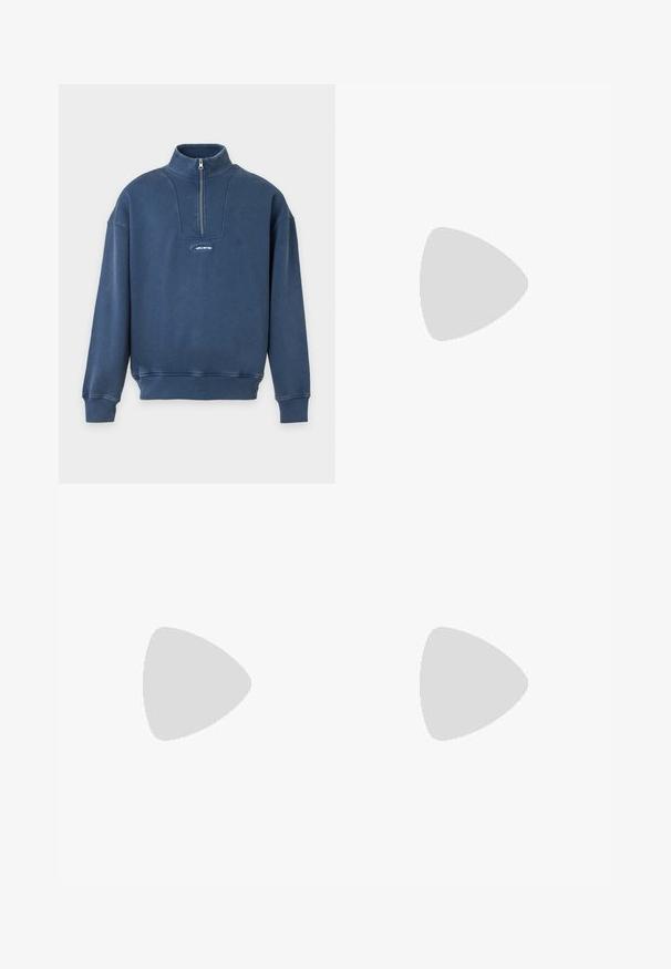 Pull bleu foncé à manches longues avec col à demi-fermeture éclair et petit logo Hollister au centre devant.; T-shirt bleu clair à manches courtes et col rond, coupe ample, présenté sur un fond blanc uni.; Pantalons larges marron en coton, avec un design classique à cinq poches, une fermeture éclair et des détails de couture visibles.; Chaussure de bateau en cuir marron avec des lacets noirs, des coutures beiges et une semelle robuste, suspendue au-dessus d'un sol en béton gris.