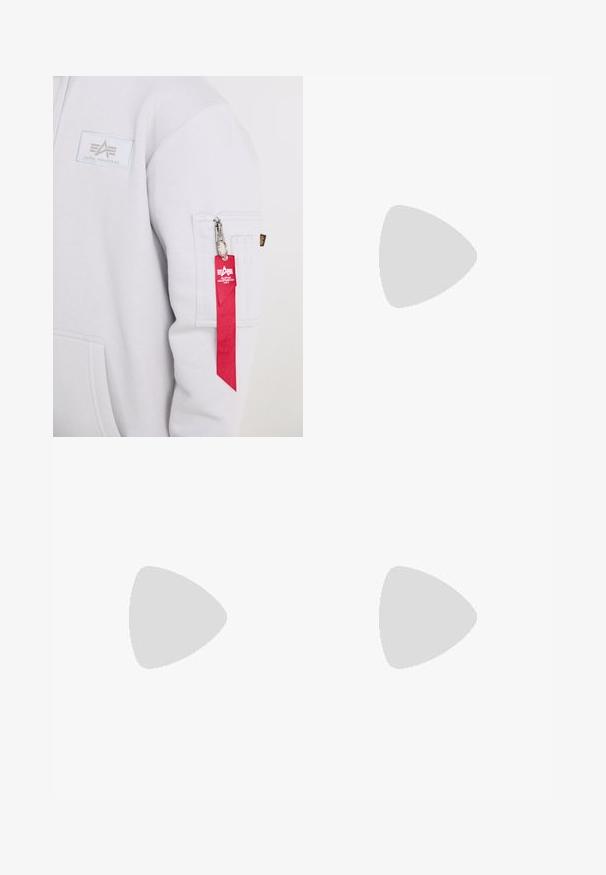 Witte mouw van een Alpha Industries sweatshirt met een zak met rits en een rood stoffen sleutelhangerlabel bevestigd aan de ritssluiting.; Wit T-shirt met kleine zwarte schets van twee personen die schilderen, één zittend en één staand, met een rode vlek op het canvas in een atelier.; Zwarte sportshorts van zachte stof, met een elastische tailleband en twee zijzakken voor gemak.; Zwarte en grijze sport sneaker met meshpanelen, vetersluiting, dikke zool en een klein krokodillologo aan de zijkant.