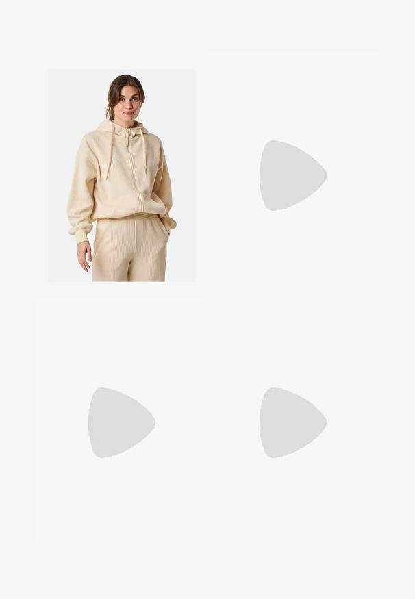 Crème ribbed hoodie met een rits aan de voorkant, grote capuchon en koordjes. Bevat een kangoeroezak en heeft een relaxed fit. Gecombineerd met ribbed broek.; Witte katoenen t-shirt met korte raglanmouwen. Heeft een opdruk van de tekst "MALDIVES" in donkergrijs, samen met logo's en de jaren 2008 en 2014.; Wijd gesneden jeans van lichtblauwe denim met vervaging, met een vijf-pocket ontwerp, knoopsluiting en minimale stikdetails.; Bruine en bordeauxkleurige sneaker van suede en mesh materiaal, met drie crèmekleurige strepen, een gemêleerd stof op de tong en een rubberen zool.