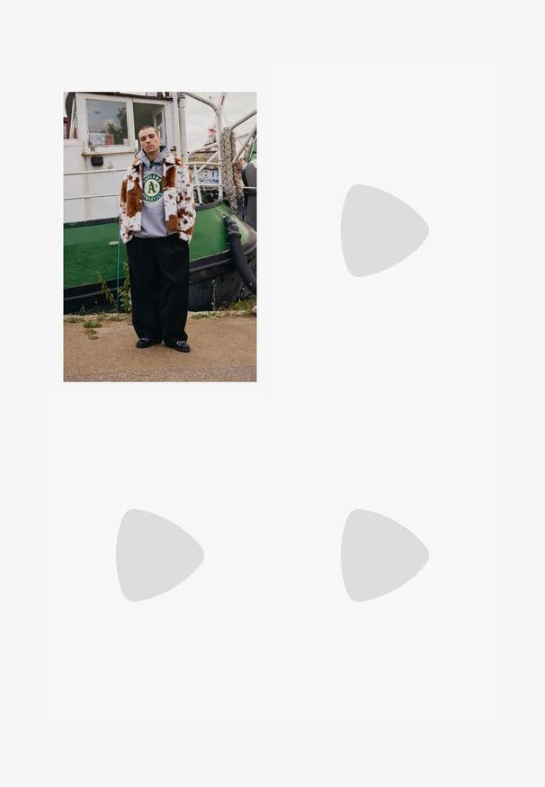 Jovem vestido com um casaco de pele castanho e branco com manchas por cima de um capuz dos Oakland Athletics, está à beira-mar, perto de um barco verde.; Camisa preta de manga longa com fecho éclair, feita de tecido leve; apresenta um colarinho redondo e um pequeno logotipo impresso no lado superior esquerdo.; Calças largas pretas com pregas na frente, bolsos laterais e uma textura de tecido suave. Projetadas para um ajuste confortável.; Botim de tornozelo em camurça bege com colarinho acolchoado preto, ilhós de metal dourado, detalhes de costura branca e sola de borracha robusta com sulcos pronunciados.