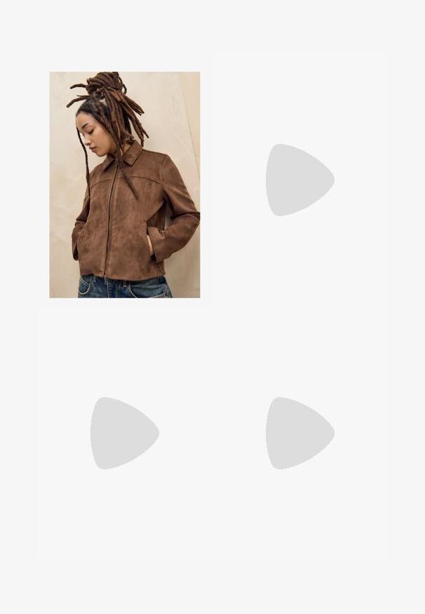 Ung person med långa bruna dreadlocks står mot en ljus trävgg iklädd en brun mockajacka och blå jeans, med händerna i fickorna och huvudet lutat ner.; Vit kortärmad blus med krage, som har en vriden knutdetalj framtill och en knappslå. Slät ribbtextur.; Breda, ljusbruna bomullshängselbyxor med hög midja, framfickor och en enda knappstängning. Slät textur, avslappnad design.; Tan mockasiner med en mjuk textur, sömnadsdetaljer och en gummisula, med en öglad häl för enkel påtagning.