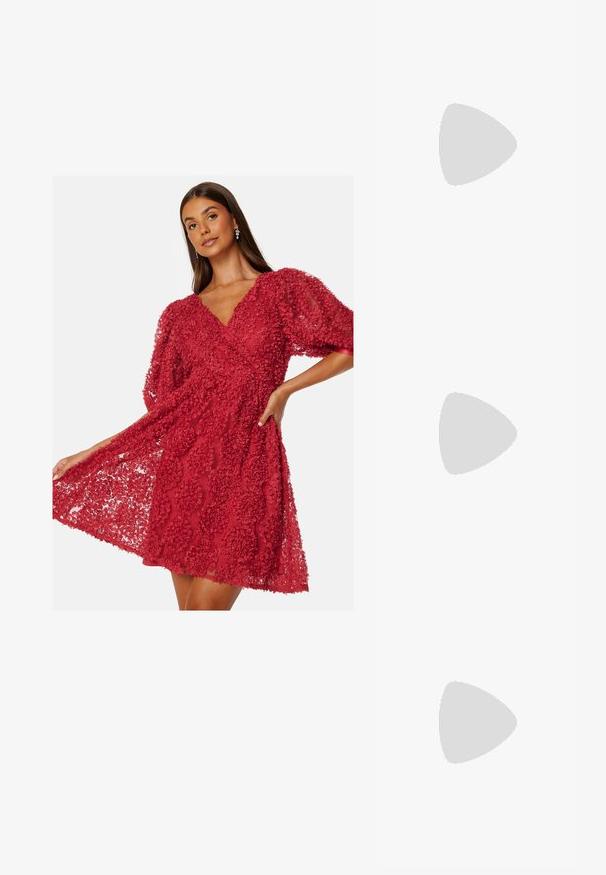 Czerwona sukienka z teksturowanej tkaniny w kwiatowy wzór. Posiada krótkie, bufiaste rękawy oraz dekolt w kształcie litery V. Rozszerzona krawędź.; Biała szpilka slingback z ostrym noskiem, gładkim materiałem wierzchnim, niskim obcasem blokowym oraz wycięciami po bokach i regulowanym paskiem z klamrą.; Okrągłe okulary przeciwsłoneczne z ciemnymi soczewkami i złotymi ramkami. Modelka nosi jasnoniebieski kardigan w prążki z guzikami i białymi rękawami w kontrastowym kolorze.; Portfel na nadgarstek marki Michael Kors w beżowej, pokrytej płótnem, z powtarzającym się wzorem logo MK, złotym okuciem i odpinanym łańcuszkowym paskiem.