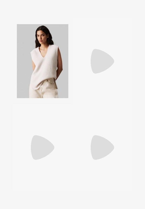 Donna con lunghi capelli scuri che indossa un maglione beige senza maniche con scollatura a V e pantaloni chiari a vita alta, in piedi contro uno sfondo grigio uniforme.; Pantaloni a gamba larga in tessuto crema. Presentano una texture liscia, una vita strutturata, cuciture frontali e nessun abbellimento visibile.; Sneakers bianchi in pelle con tre strisce nere e ricami floreali in viola e giallo. Suola in gomma marrone. Abbinati a calze a coste beige.; Occhiali da sole rotondi con motivo tartarugato e lenti trasparenti. Presentano una montatura spessa e leggera con steli sottili.