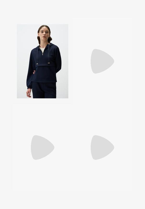 Marineblaue Kapuzenjacke aus weichem Stoff; mit durchgehendem Reißverschluss, goldenen Hardware-Akzenten und elastischen Bündchen für eine bequeme Passform.; Weit geschnittene Hose aus leichtem beigen Baumwollstoff mit Knopfverschluss, dezent fransigem Saum und lockerem Schnitt.; Beige Wildleder-Ankle-Boot mit Plateausohle, abgerundetem Zehenbereich und abgesteppten Akzenten; verfügt über eine Zuglasche hinten für einfaches An- und Ausziehen.; Schildpatt-Sonnenbrille mit grünen Gläsern, rechteckigem Rahmen und auffälligem Brückendesign. Besitzt dezente Markenkennzeichnung am Bügel.